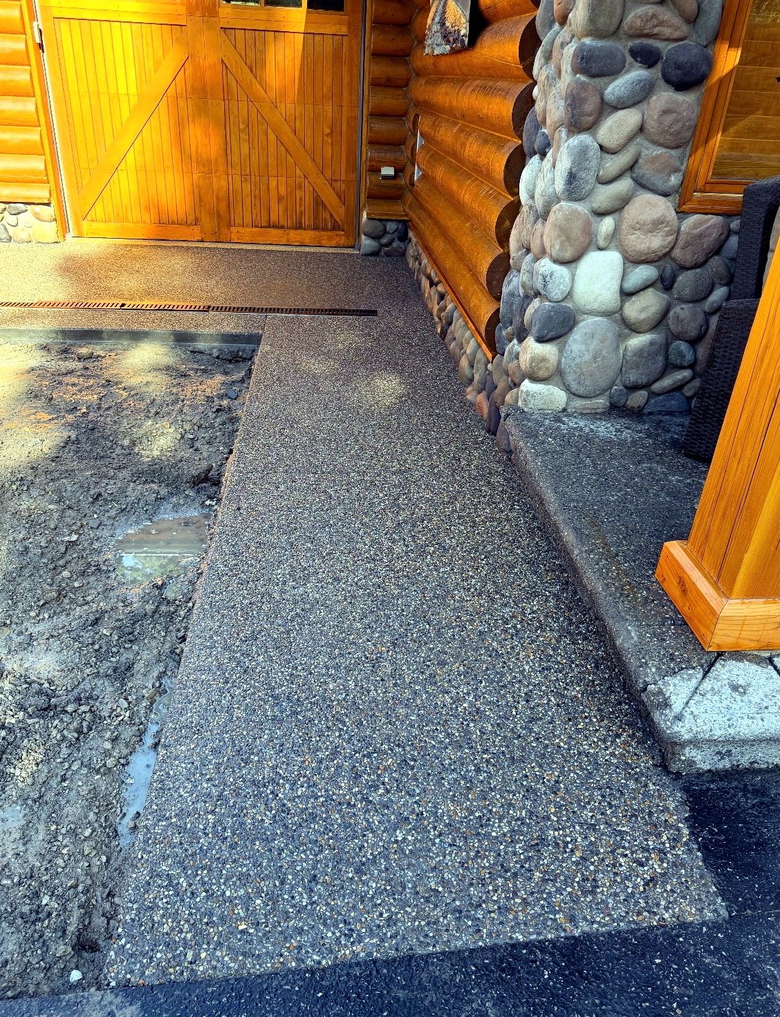 Exposed aggregate concrete walkway with integrated trench drain alongside a log home with river rock pillars, installed by SiteWorx Concrete Calgary