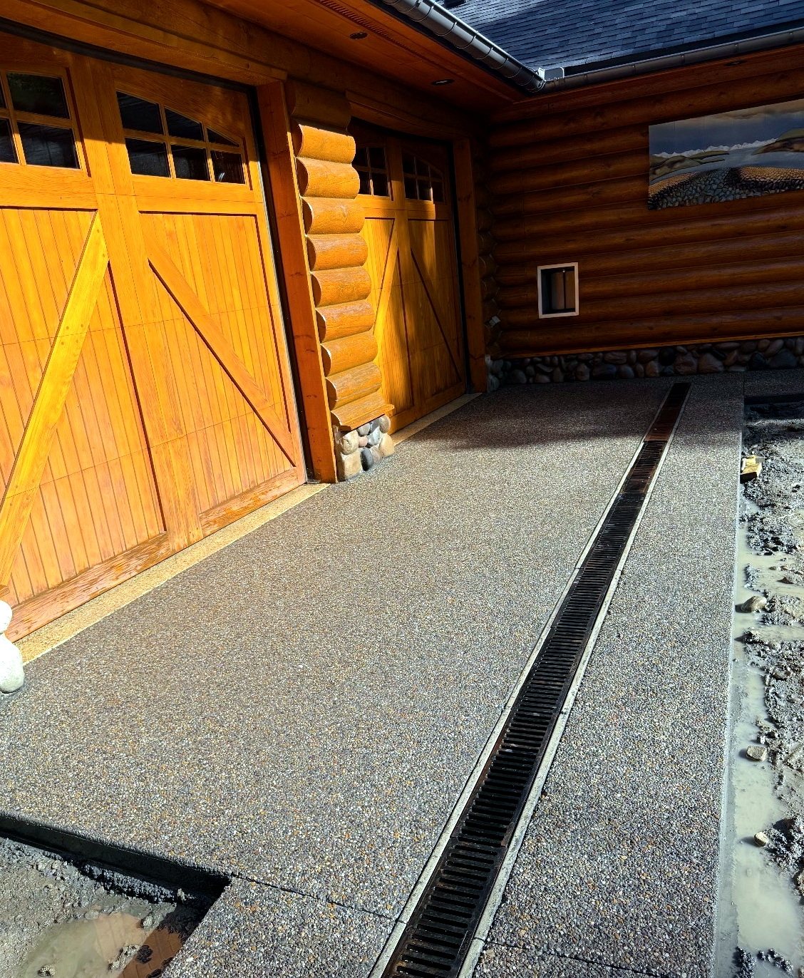 Exposed aggregate concrete driveway with a full-length trench drain channel in front of a log home garage, installed by SiteWorx Concrete Calgary